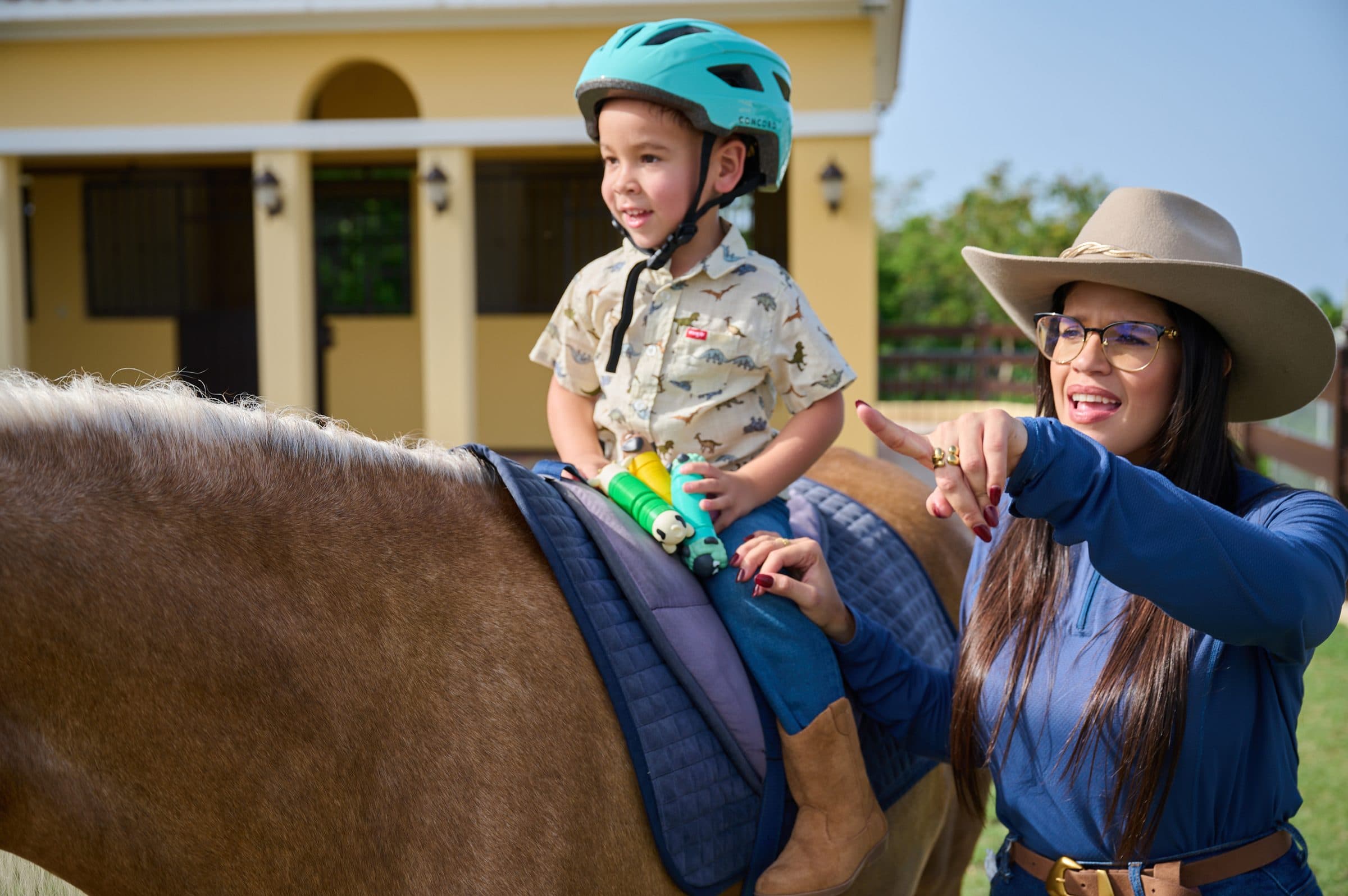 Niño sonriente con casco turquesa montado a caballo frente a las caballerizas
